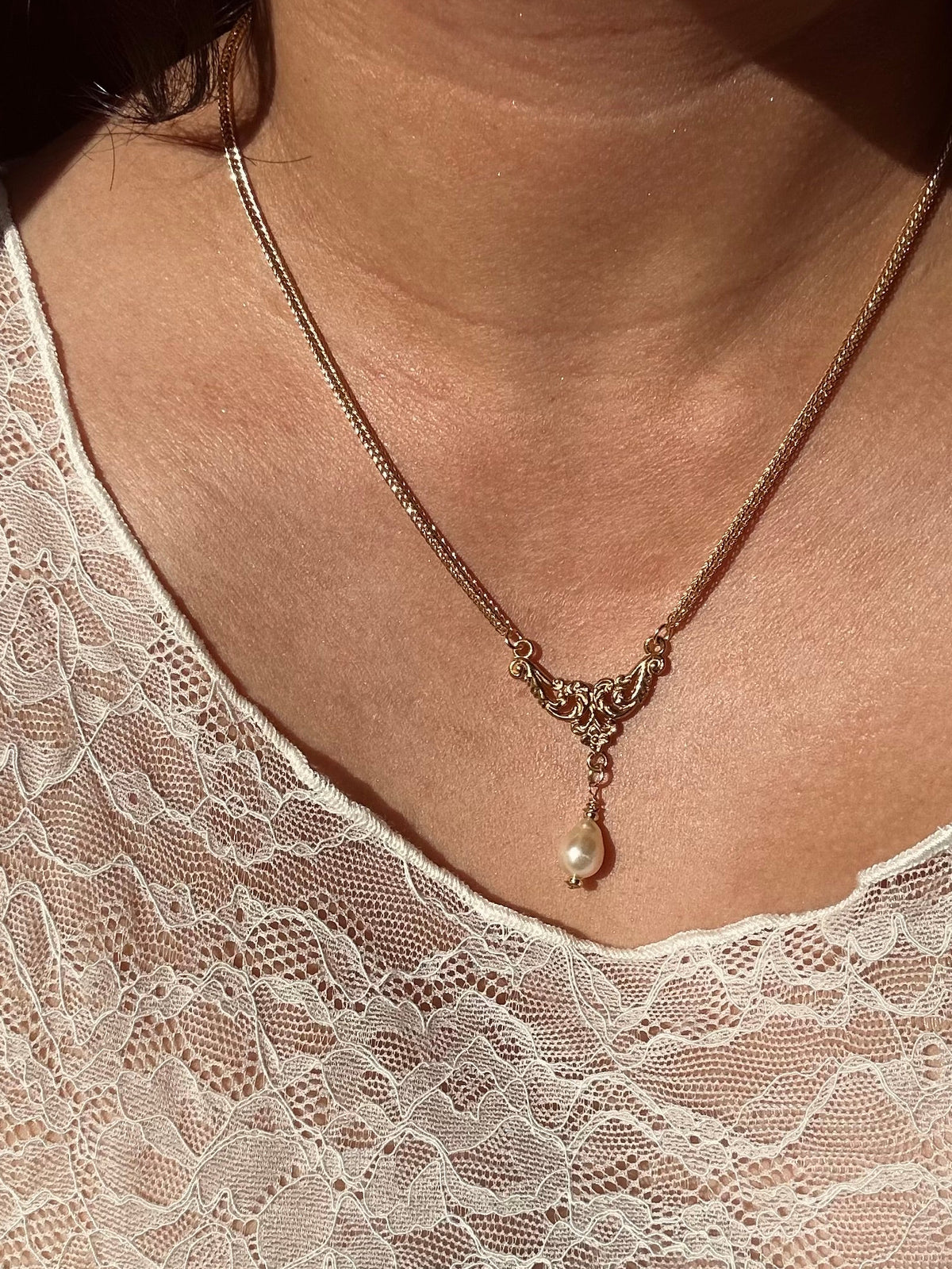 A close-up of a person wearing a delicate gold chain necklace with an ornate filigree-style center and a single pearl drop pendant, paired with a white lace top.