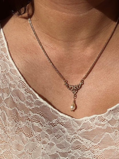 A close-up of a person wearing a delicate gold chain necklace with an ornate filigree-style center and a single pearl drop pendant, paired with a white lace top.